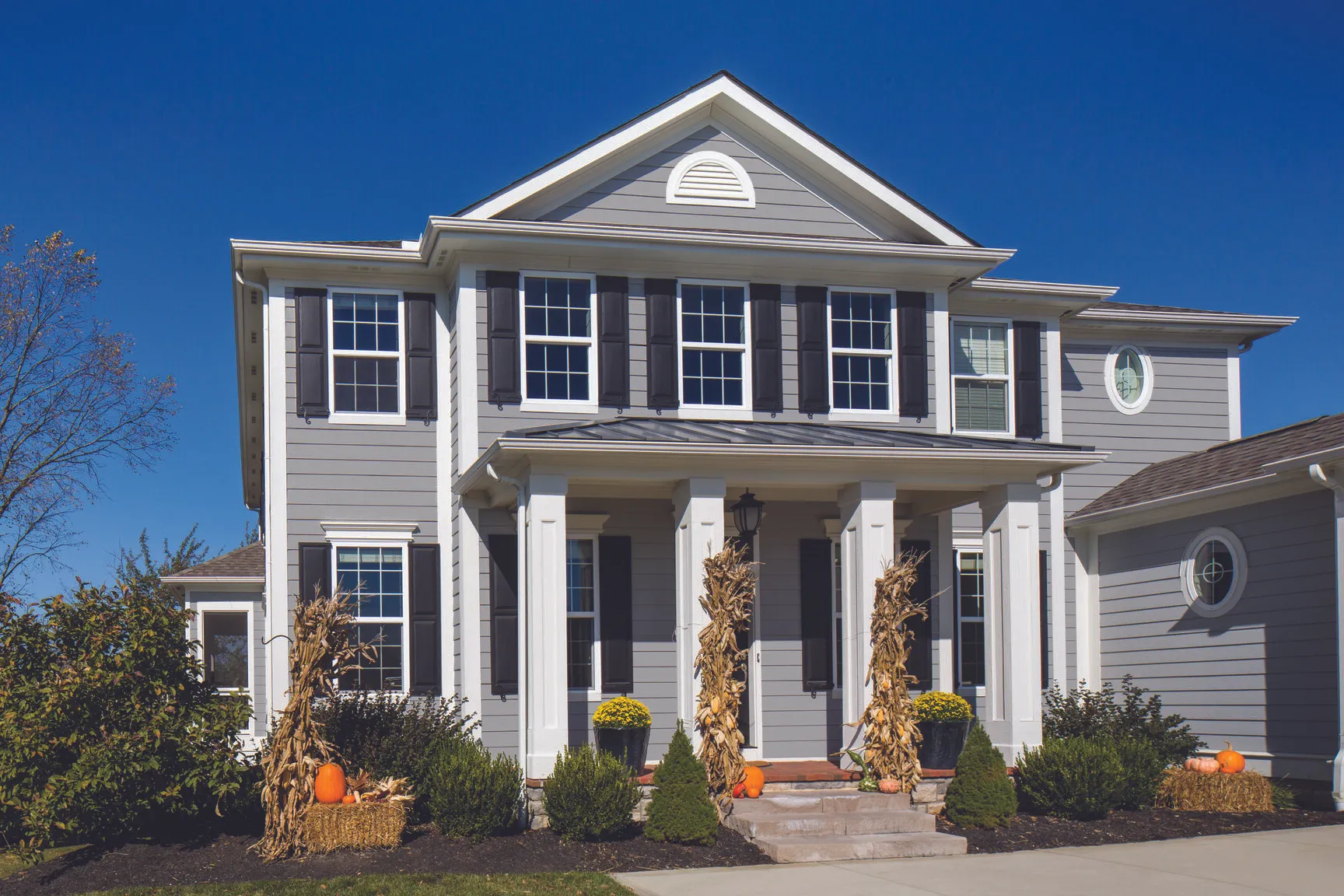 Two-story gray house with white trim, black shutters, and fall decorations including pumpkins and dried corn stalks on the front porch.
